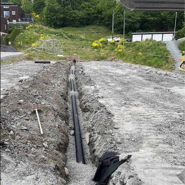 Grøft med svart dreneringsrør lagt i grus på byggeplass.