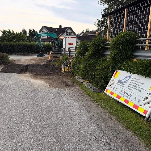 Gravemaskin rydder vei i boligområde med firmaskilt langs veien.