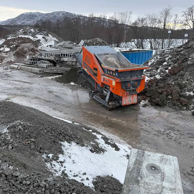 Knusemaskin på anleggsplass med betongelementer og snødekte fjell i bakgrunnen.
