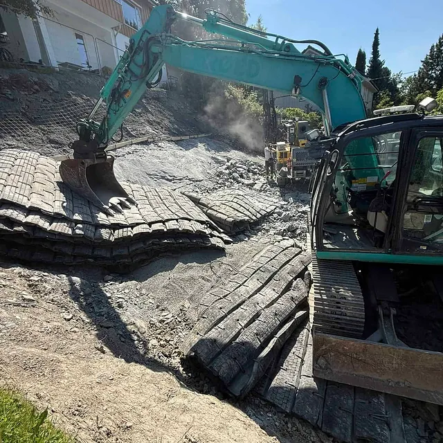 Gravemaskin meisler fjell ved skråning foran bolighus.