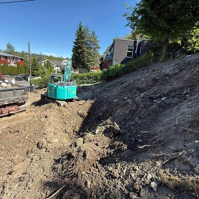 Gravemaskin laster stein i lastebil ved skrå tomt i boligfelt.