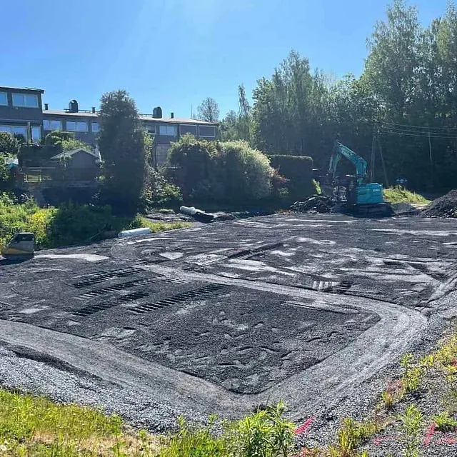 Planert tomt med grusdekke, gravemaskin og rekkehus i bakgrunnen.